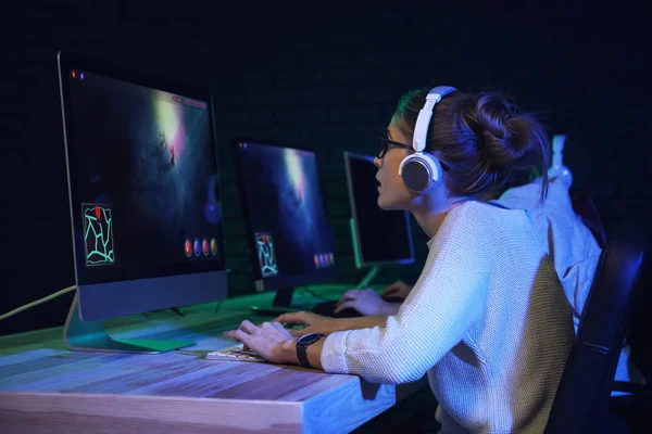 A player in a headset leaning toward a monitor at a station, the screen glow lighting a row of displays