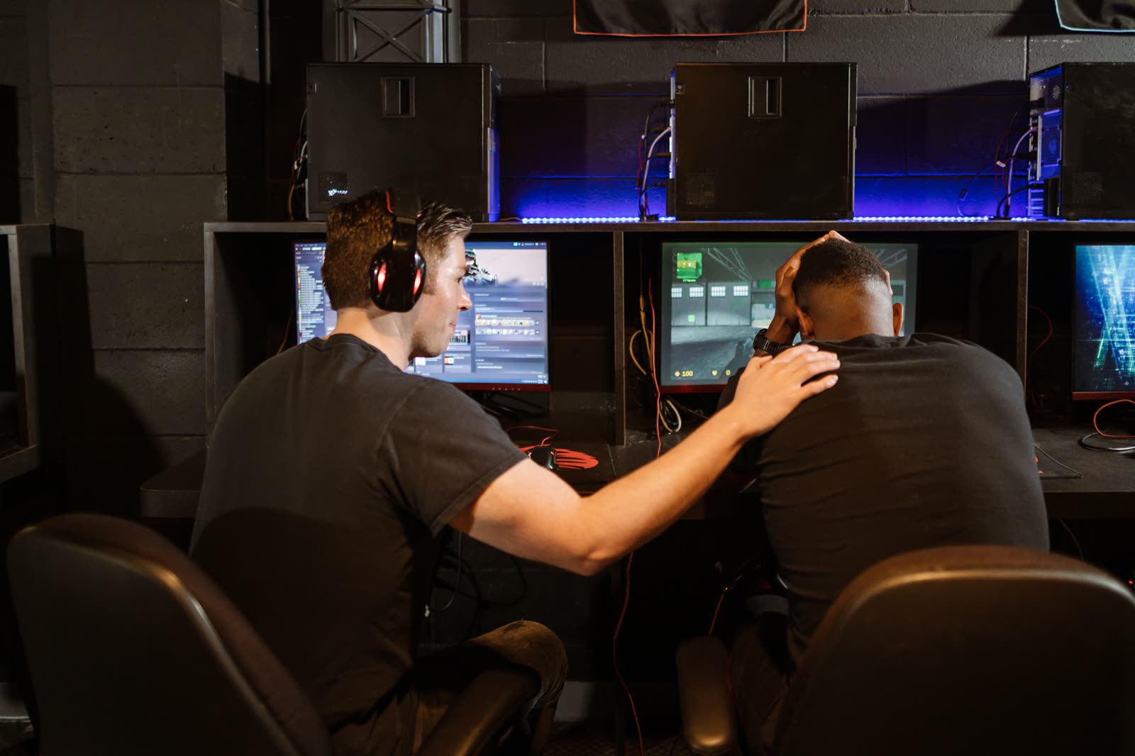 Two players seated side by side at a lit station row, one leaning over to coach the other